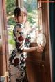 A woman in a cheongsam standing by a window holding a fan.