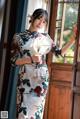 A woman in a cheongsam standing by a window holding a fan.